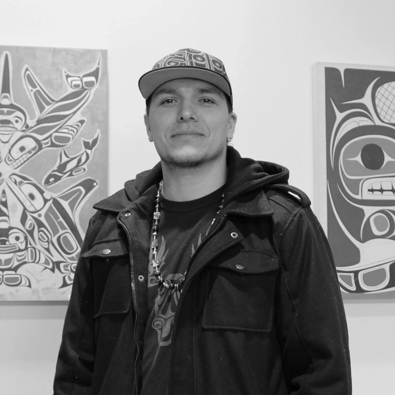 Black and white photo or Haisla, Heiltsuk artist Paul Windsor smiling in front of Indigenous art work