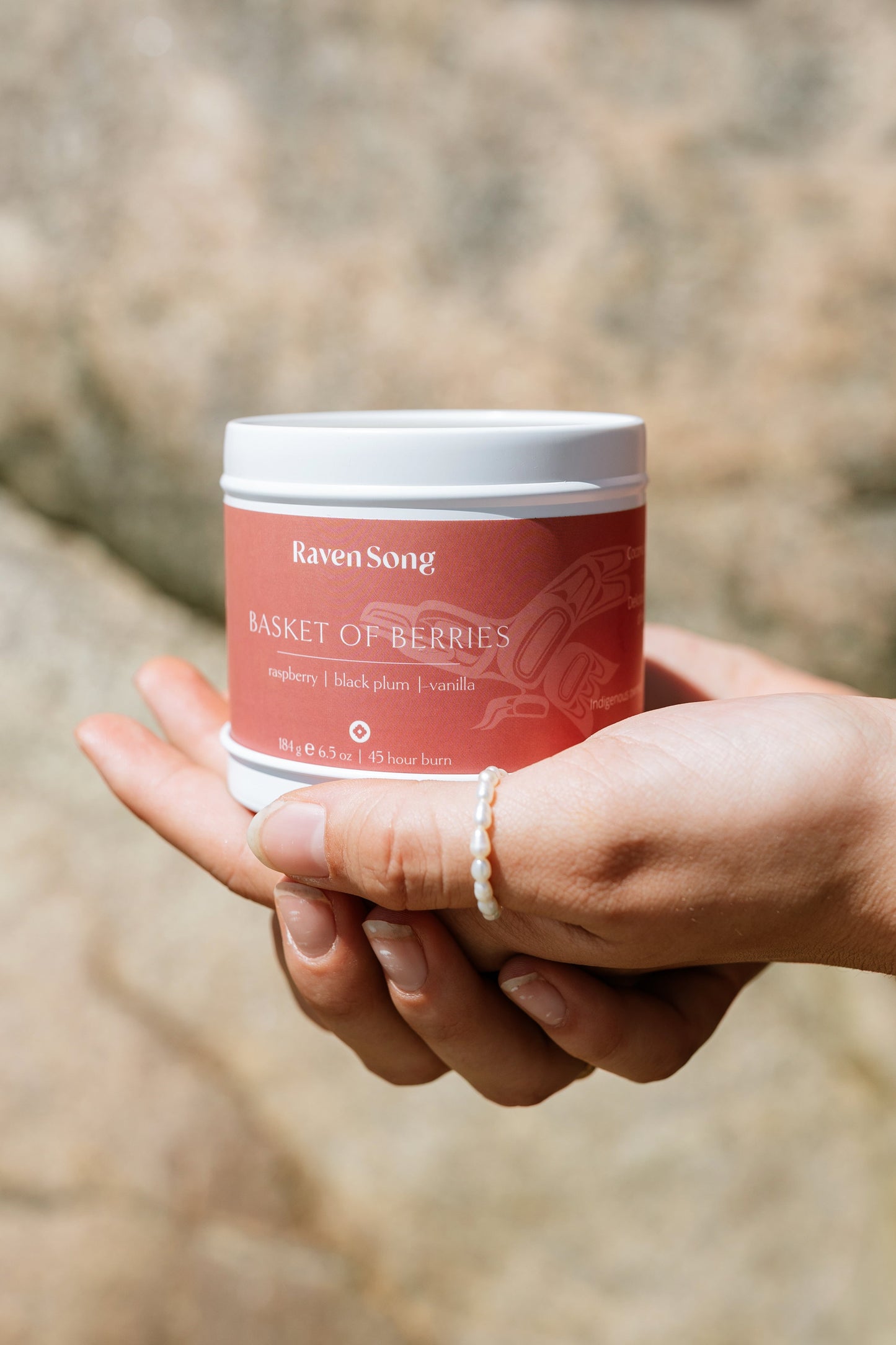 Hand holding a red candle jar labeled 'Raven Song Basket of Berries' against a blurred natural background