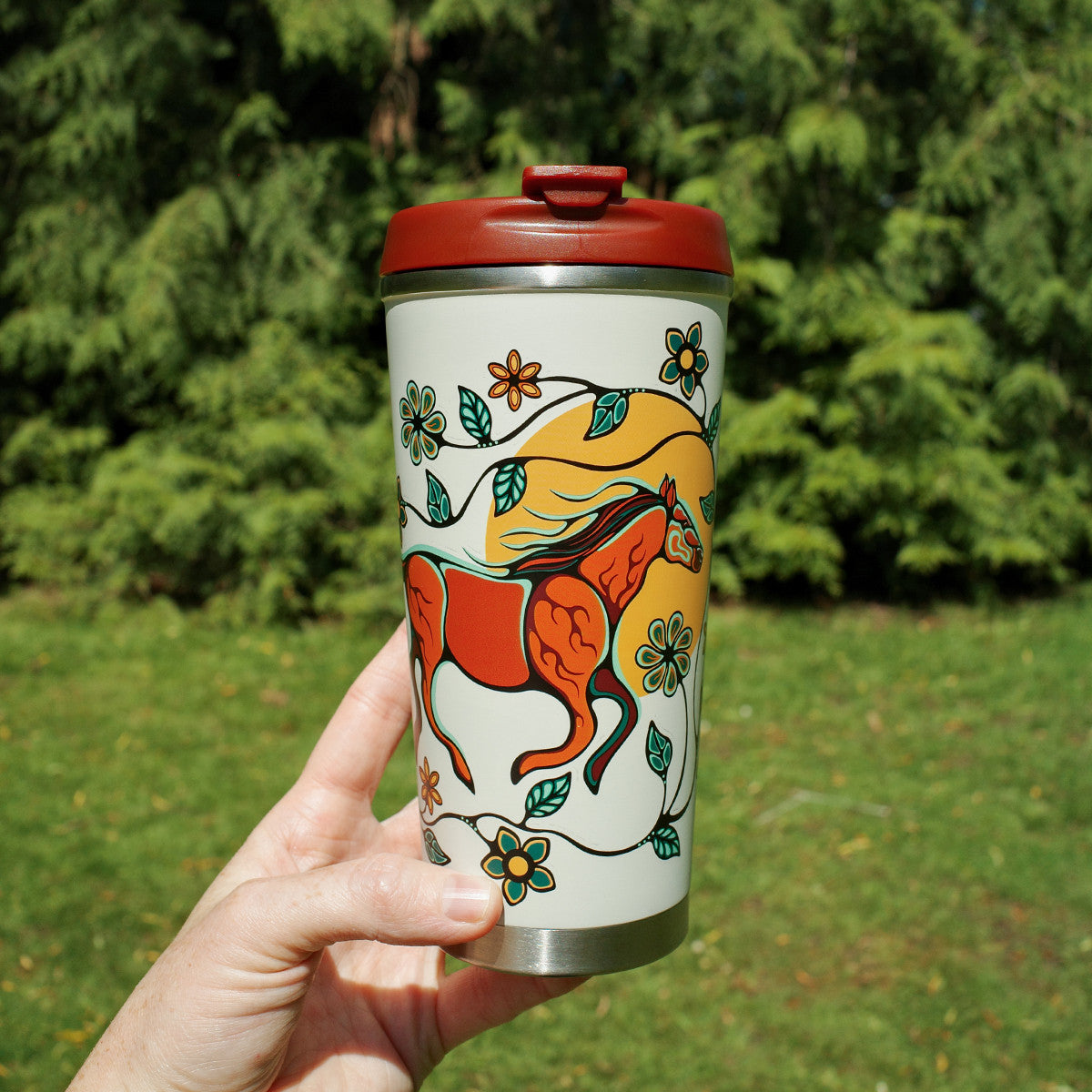 Hand holding a colorful travel mug with a fox design against a green outdoor background
