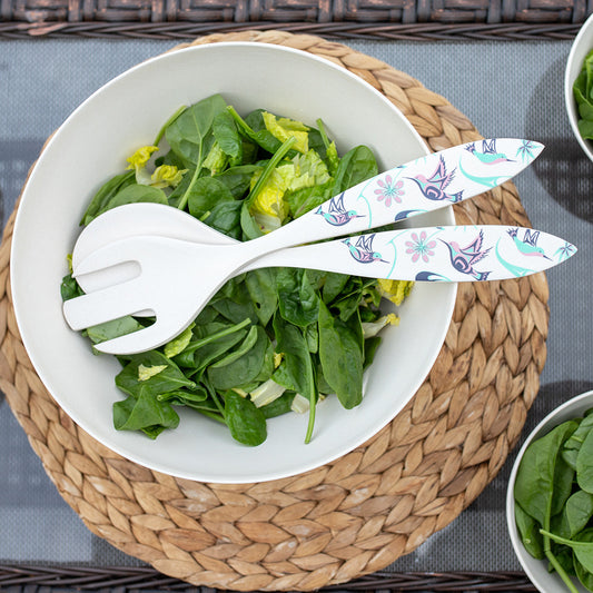 White bowl with green salad and decorative salad servers on a woven placemat.