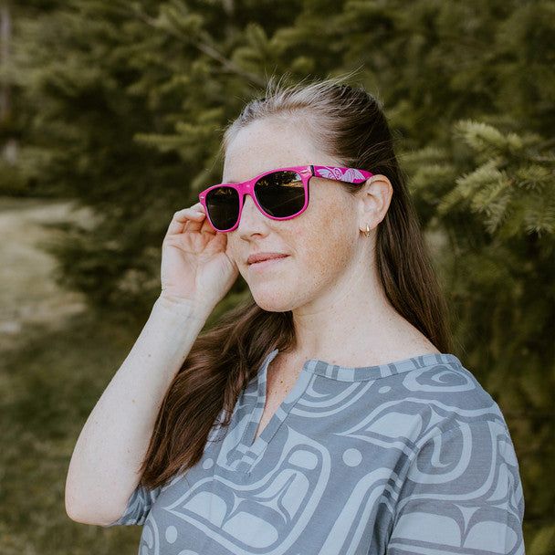 Woman wearing the pink Hummingbird sunglasses standing in front of a cedar tree