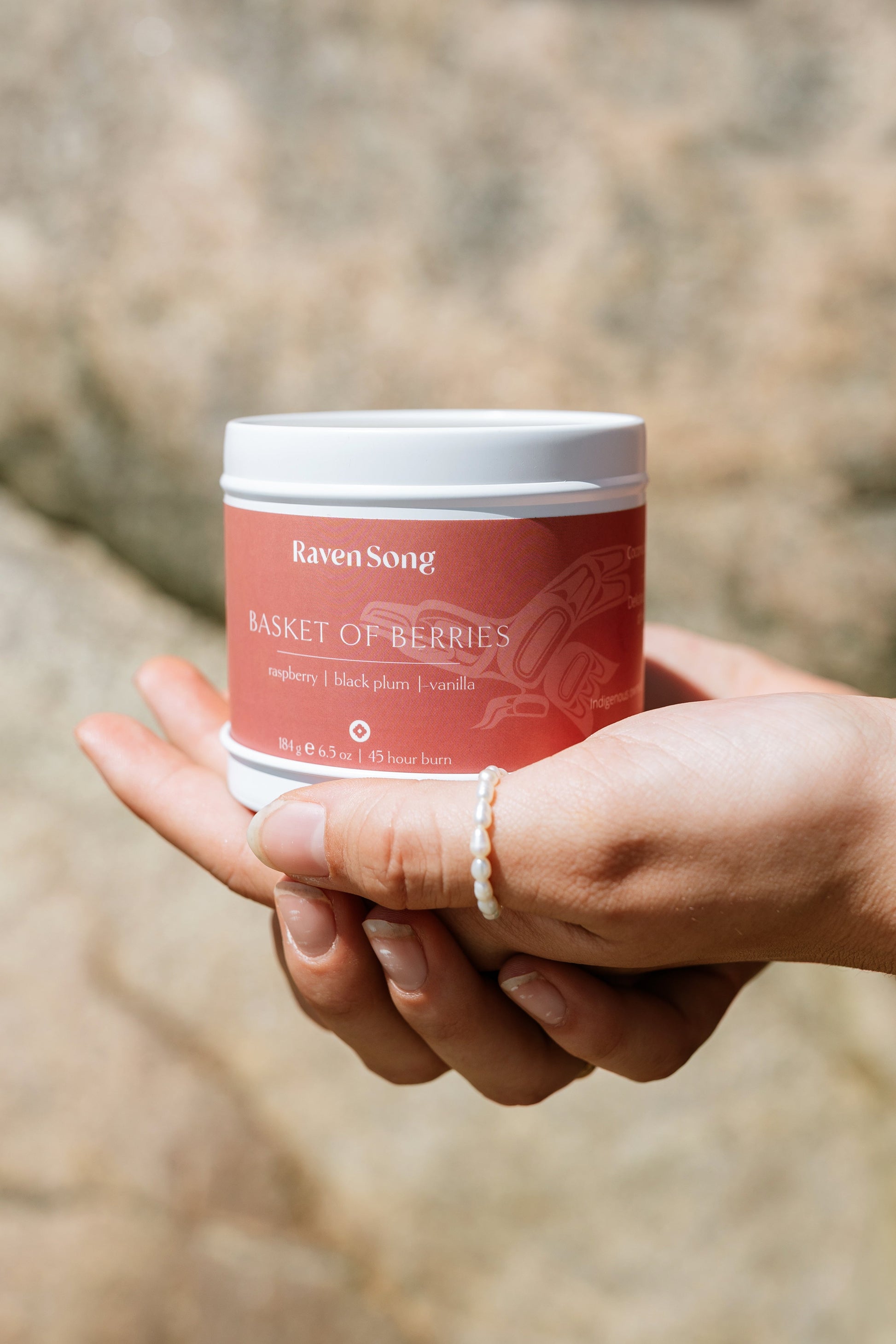 Hand holding a red candle jar labeled 'Raven Song Basket of Berries' against a blurred natural background