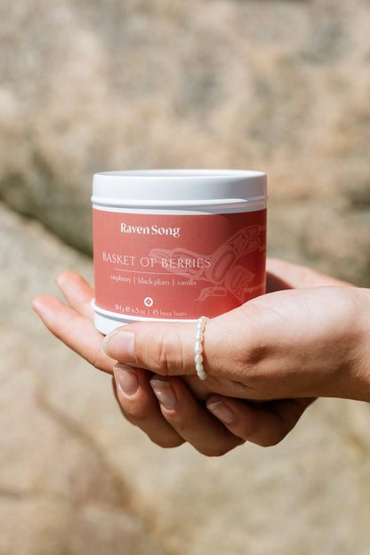 Hand holding a red candle jar labeled 'Raven Song Basket of Berries' against a blurred natural background
