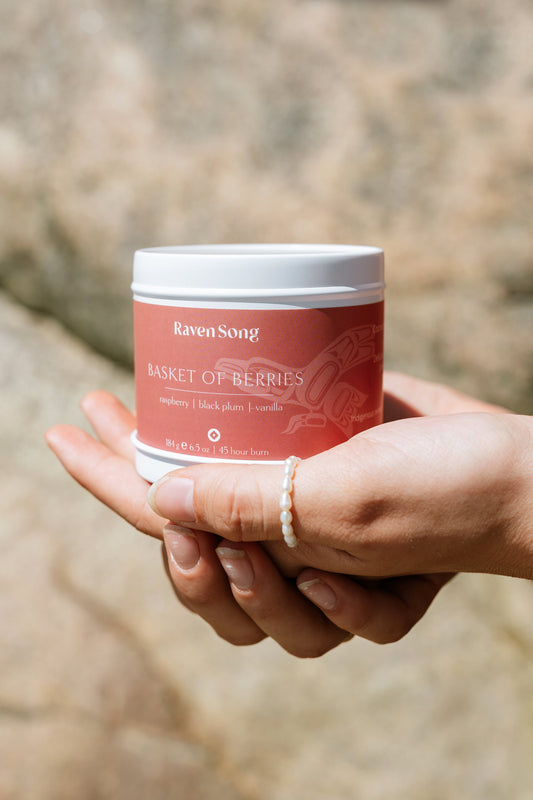 Hand holding a red candle jar labeled 'Raven Song Basket of Berries' against a blurred natural background