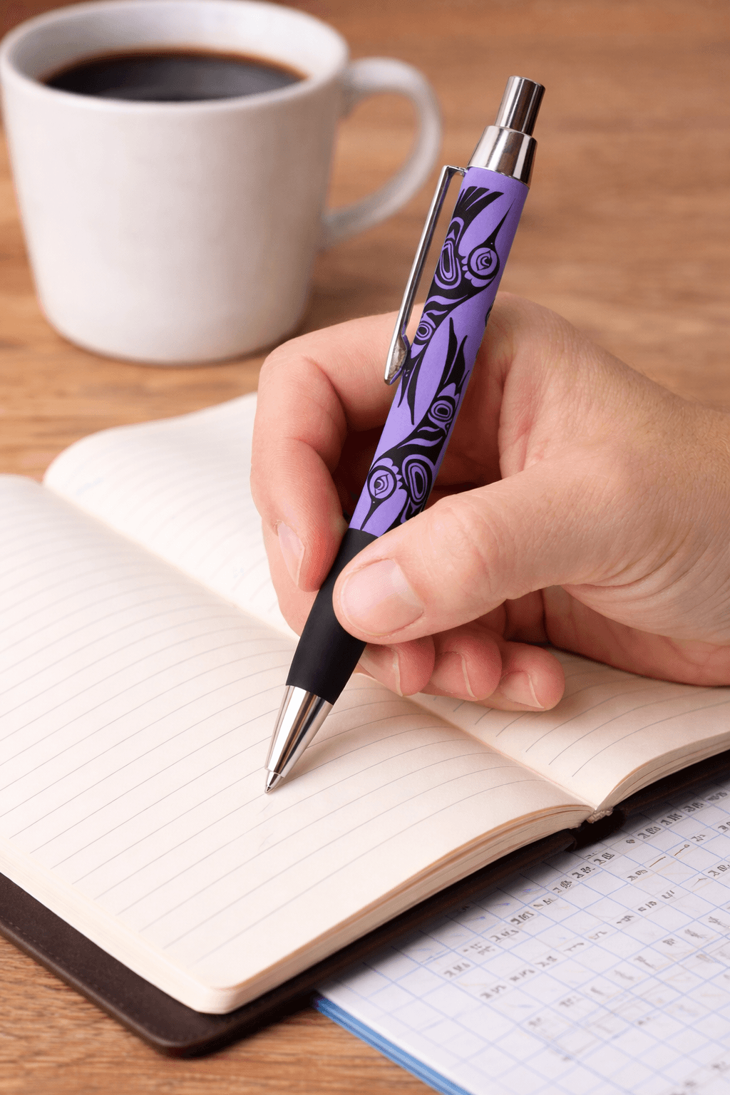 Pen with indigenous artwork of hummingbird by Paul Windsor. Photo shows a persons hand using the pen to write in a notebook on a desk. There is a coffee in the background
