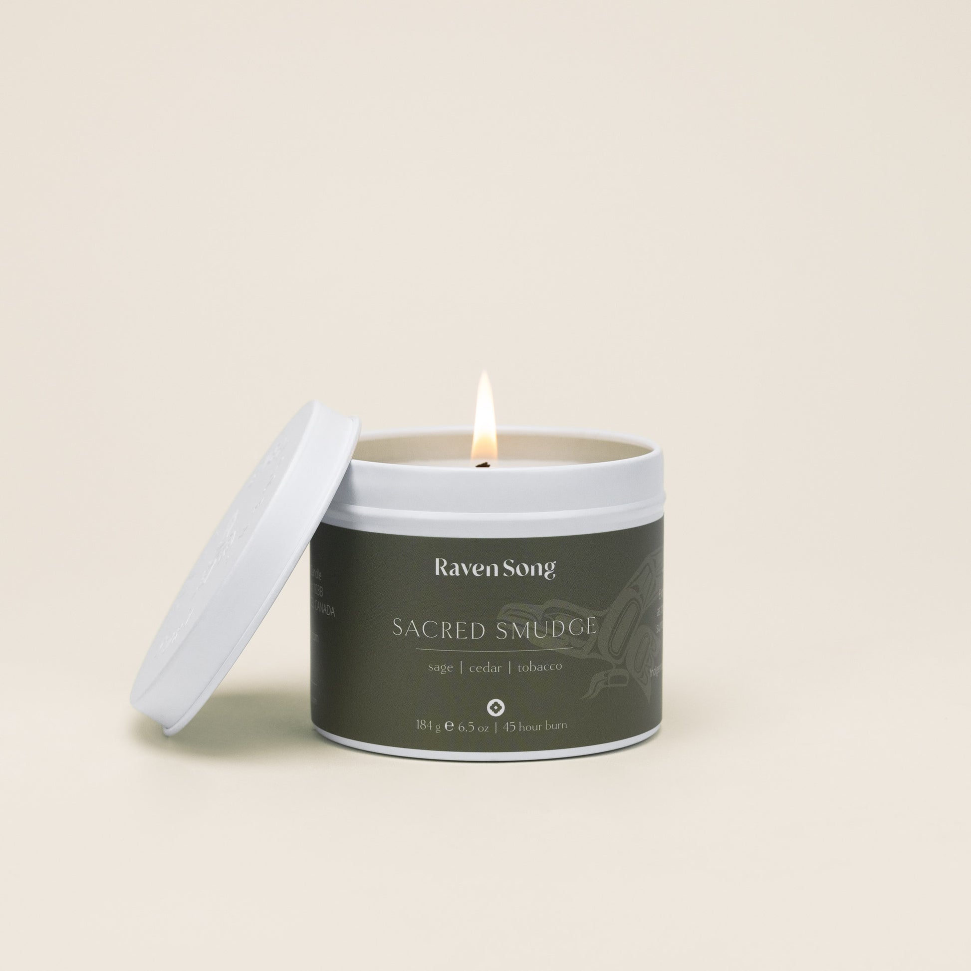 Candle in a container labeled 'Raven Song Sacred Smudge' with a burning wick