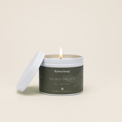Candle in a container labeled 'Raven Song Sacred Smudge' with a burning wick