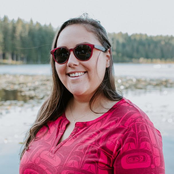 Woman wearing Eagle Vision sunglasses by Haida artist Allan Weir, standing on a beach with trees in the background
