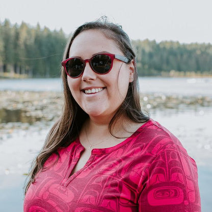 Woman wearing Eagle Vision sunglasses by Haida artist Allan Weir, standing on a beach with trees in the background