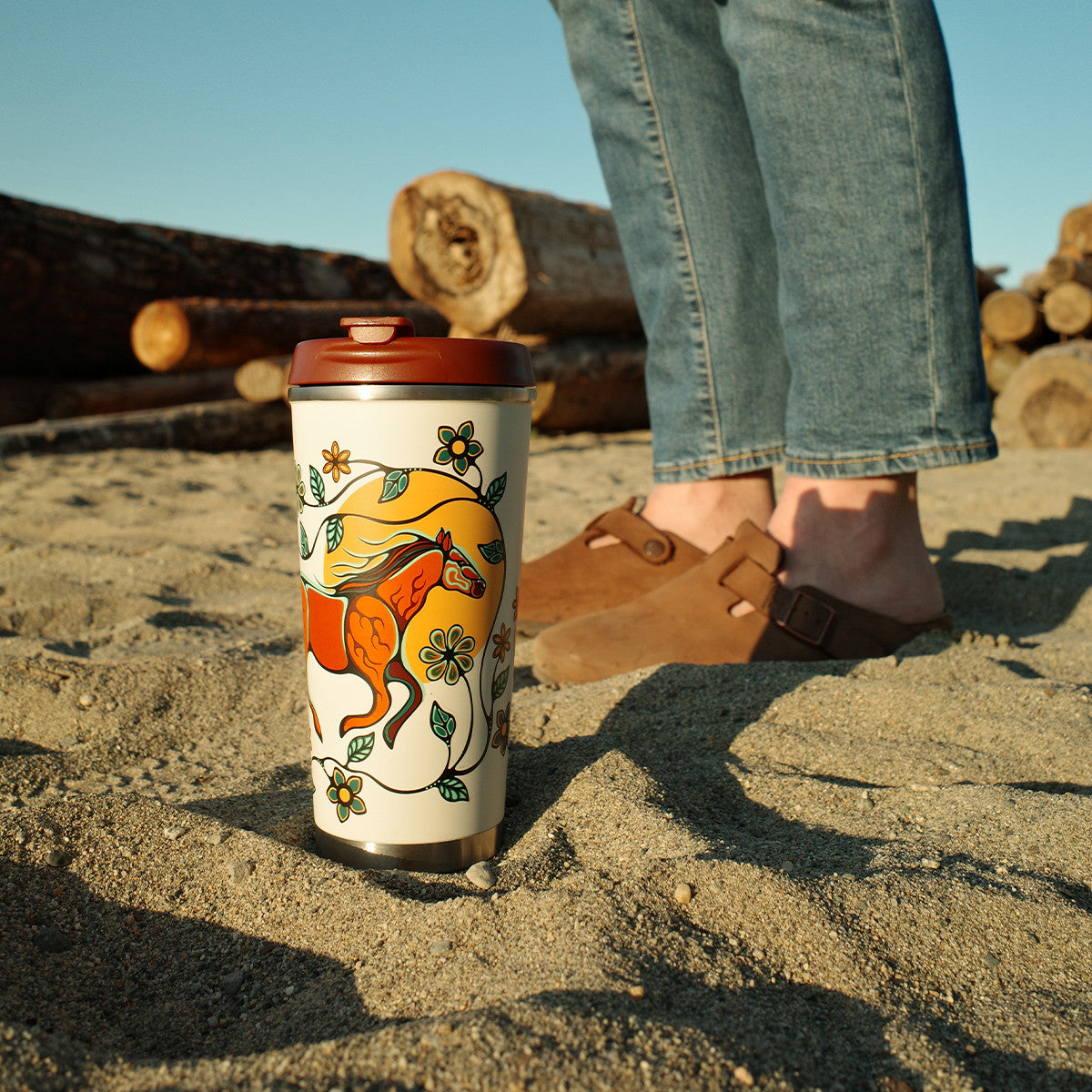 Person standing on a sandy beach with a decorated cup and wooden logs in the background