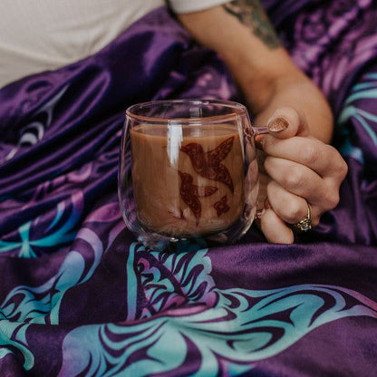 Women holding Double walled Glass Mug with Indigenous Design Hummingbird By Simone Diamond with purple blanket underneath