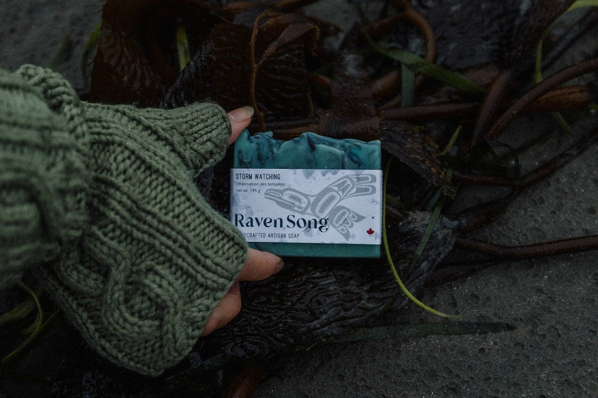Storm Watching soap by RavenSong in its wrapped label showing net weight 115 g, held above beach kelp