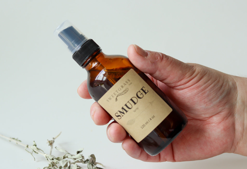 Hand holding the 4 oz Sweetgrass Soap Smudge Spray amber bottle, dried sage below.
