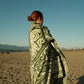 Person wrapped in a green and white patterned blanket on a beach with mountains and city skyline in the background.